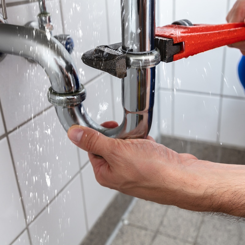 Plombier utilisant une clé serre-tube rouge pour réparer une fuite d'eau importante sur un siphon chromé sous un lavabo.