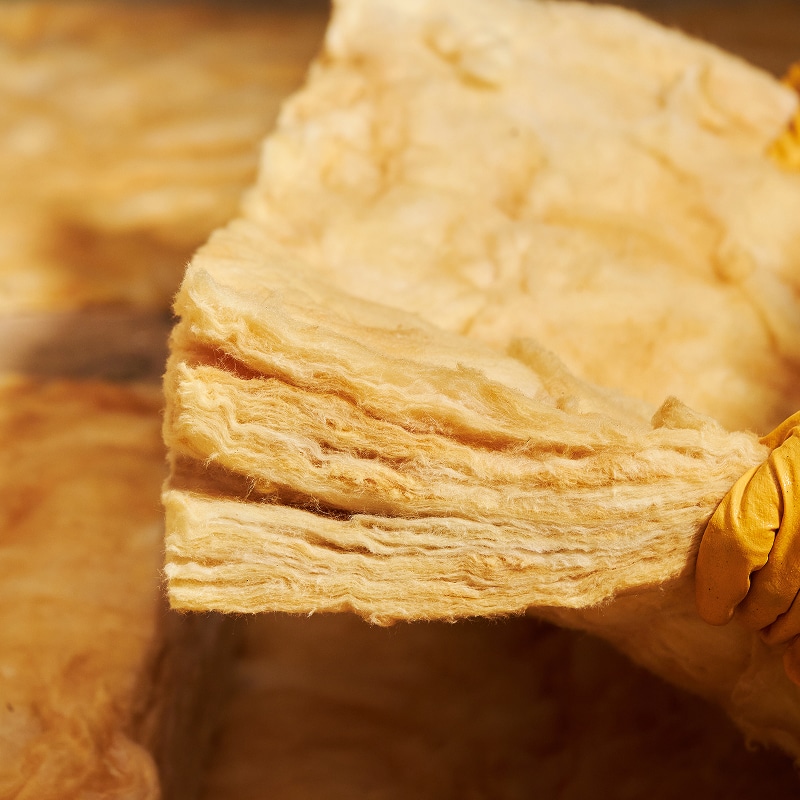 Gros plan sur un isolant thermique en laine de verre ou roche, de couleur jaune-beige, tenu par une main portant un gant de protection jaune.