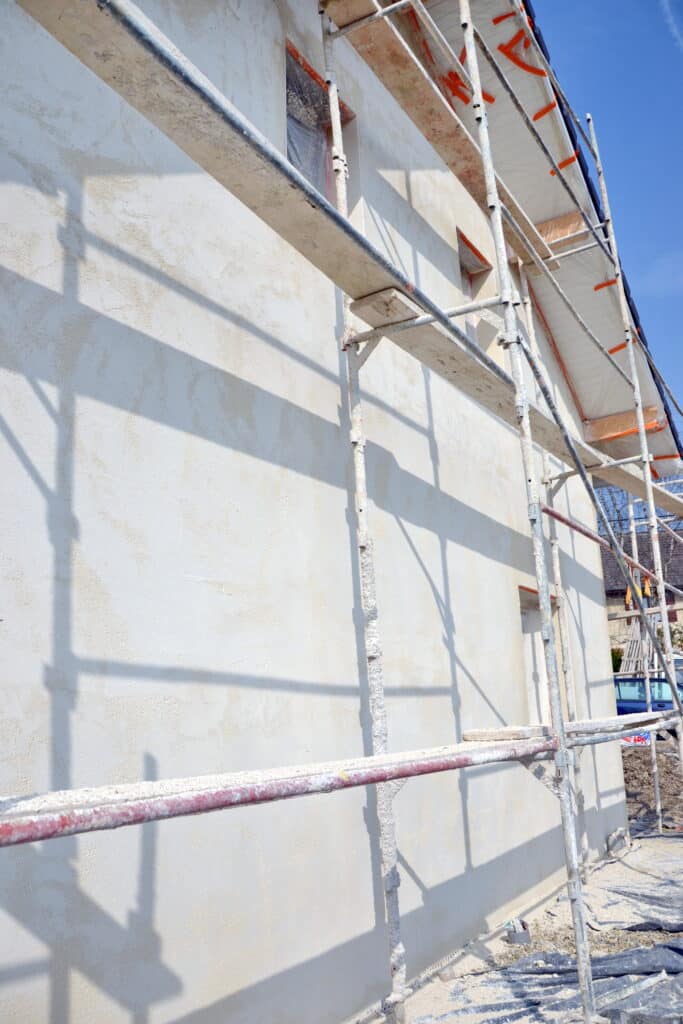 Chantier maison enduit neuf et échafaudage Façade d'une maison neuve avec enduit clair fraîchement appliqué, échafaudage métallique et ombres portées sous un ciel bleu vif.